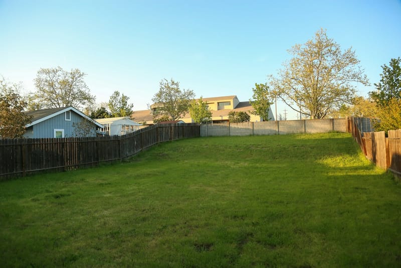 A green backyard lawn bordered by a block wall and wooden fence, with neighboring houses beyond
